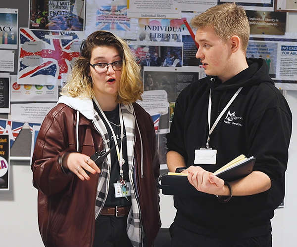 2 people talking, one is holding a walkie talkie, the other is holding a note book