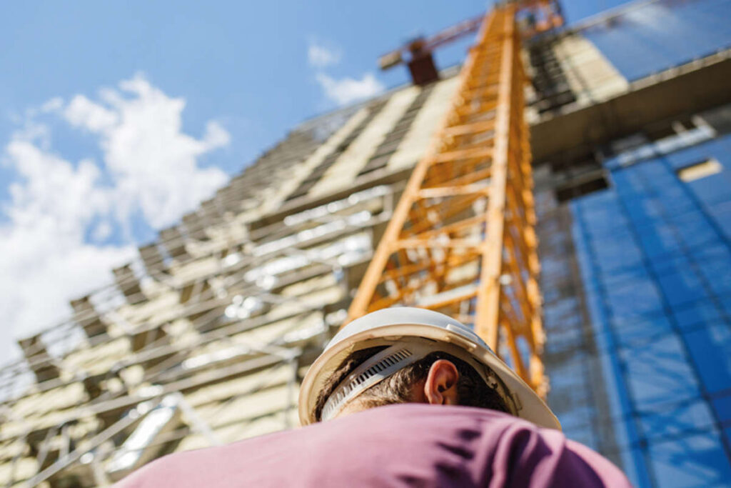 An upward shot the back of someone wearing a hard hat standing in front of a crane