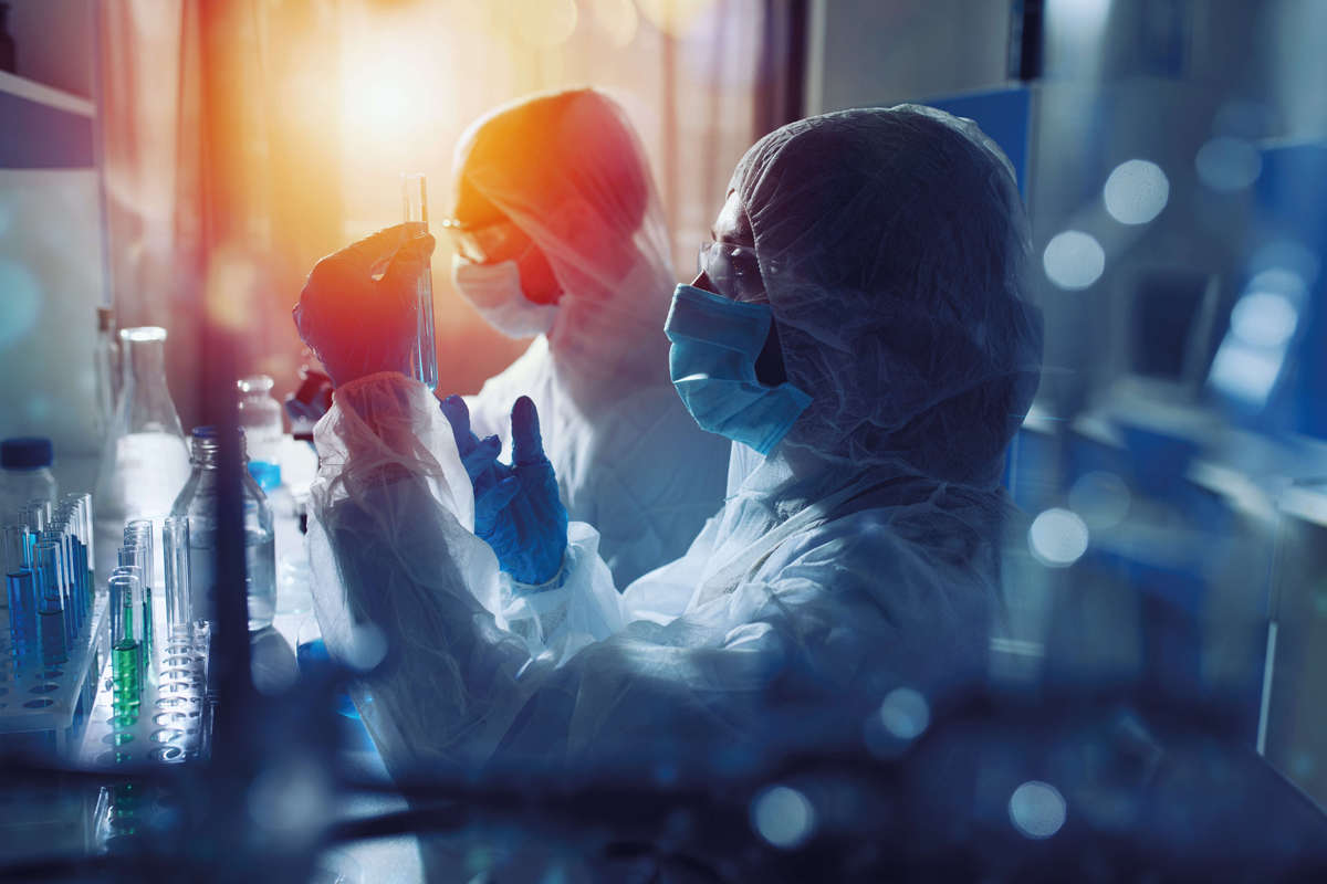 2 people in science lab, wearing white overalls, gloves, masks and safety glasses inspect blue liquid in a test tube