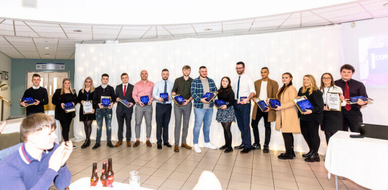 A group of apprenticeship students line up against a wall holding their awards